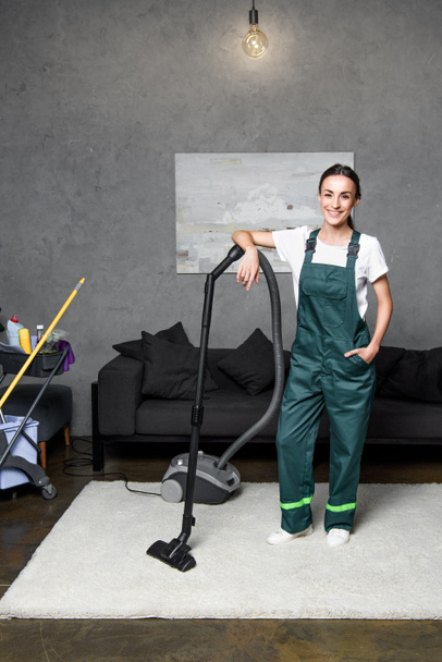Elektrikli süpürge eğilerek ve kameraya gülümseyen mutlu genç kadın temizlik şirketi işçisi  - Fotoğraf, Görsel