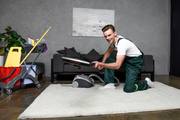 handsome young man using vacuum cleaner and smiling at camera, professional carpet cleaning - Photo, Image
