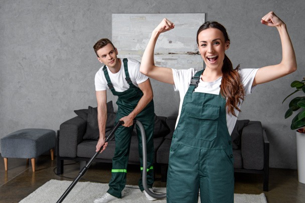 young male cleaner using vacuum cleaner and looking at female coworker showing muscles and smiling at camera - Fotoğraf, Görsel