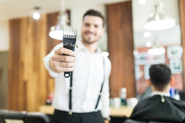 Jovem cabeleireiro masculino segurando aparador com foco na mão no salão
 - Foto, Imagem