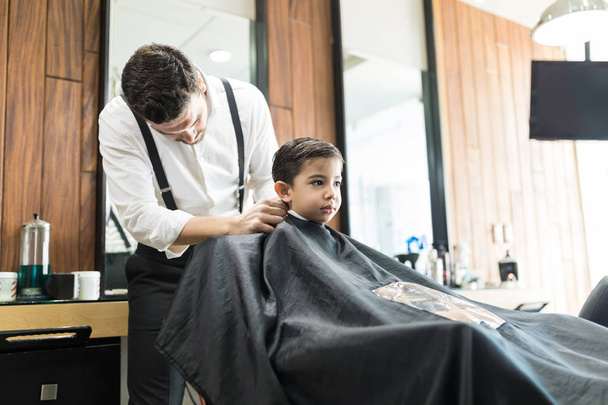 Молодой парикмахер натягивает плащ на маленького мальчика, сидящего в магазине
 - Фото, изображение