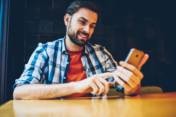 Cheerful bearded young man dressed in casual outfit reading funny news in blog on smartphone device connected to 4G internet.Positive hipster guy watching video online on modern mobile phone - Photo, Image