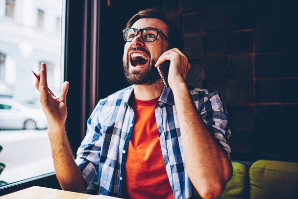 Casual dressed angry bearded young man crying and arguing during phone conversation on smartphone device.Annoyed hipster guy in eyeglasses shouting caling on modern mobile phone sitting in coworking - Foto, Imagen