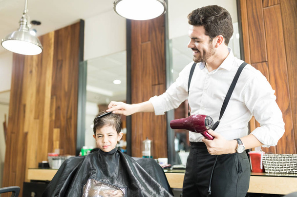 Улыбающийся молодой парикмахер расчесывает волосы мальчика в парикмахерской
 - Фото, изображение