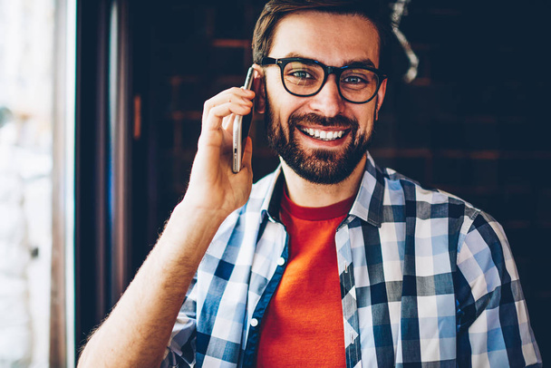 Half length portrait of successful young man in eyeglasses for vision correction smiling at camera while calling on smartphone.Positive hipster guy talking with operator about mobie benefits on phone - Valokuva, kuva