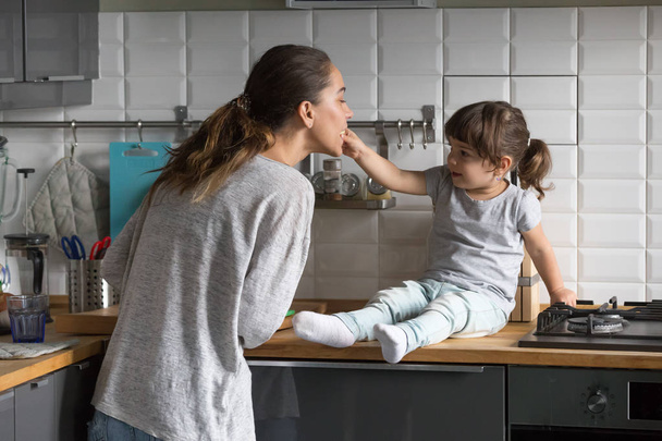 Little daughter feeding mother having fun in kitchen at home - Zdjęcie, obraz