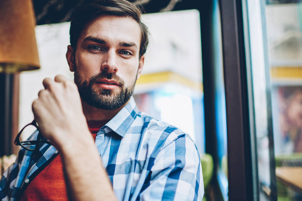 Half length portrait of handsome bearded young man looking at camera while holding eyeglasses in hands resting in coffee shop interior.Good looking hipster guy in stylish shirt sitting in cafe - Foto, Bild