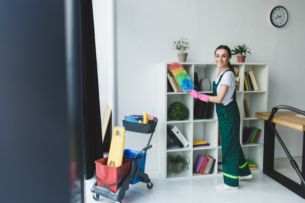 young cleaner holding duster and smiling at camera while cleaning shelves in office  - Photo, Image