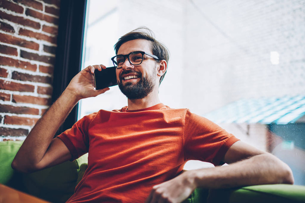 Positive hipster guy with beard calling on smartphone device to arrange meeting with friend.Joyful young man in stylish eyeglasses talking on modern mobile phone resting in coffee shop interior - Foto, Imagen