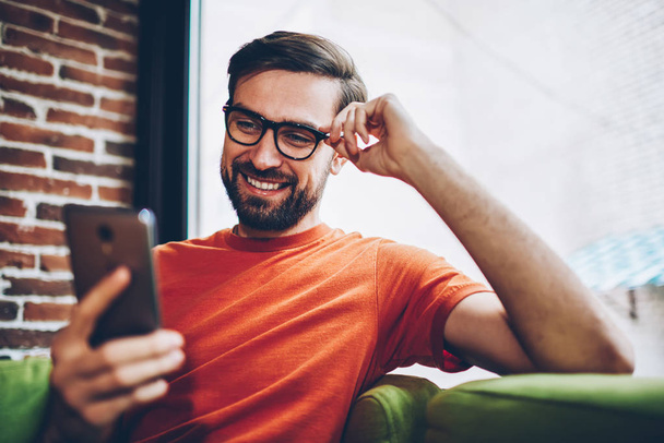 Cheerful bearded male blogger laughing while chatting with friends in social networks on modern smartphone.Positive young man watching video on cellular connected to free 4G internet - Фото, зображення