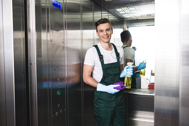 schöner junger Hausmeister mit Sprühflasche mit Waschmittel und Lappen, der im Fahrstuhl in die Kamera lächelt - Foto, Bild
