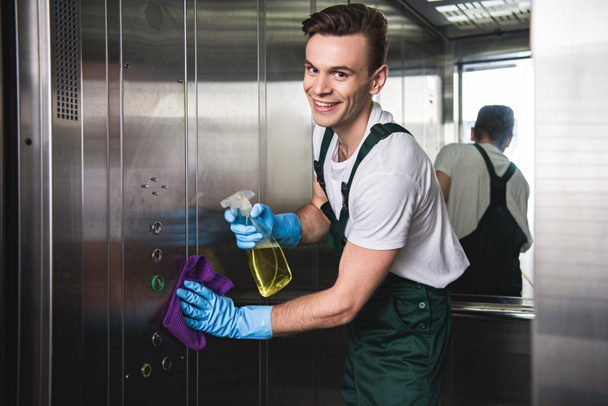 fiatal takarítás társaság-munkás tisztító lift és mosolyogva kamera    - Fotó, kép