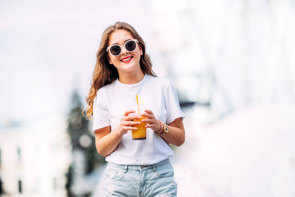 Portrait of wonderful white female model with bright makeup expressing energy in good day.Adorable girl in short denim shorts and bright t-shirts, sunglassesen joying festive in amusement park. holds cocktail - 写真・画像