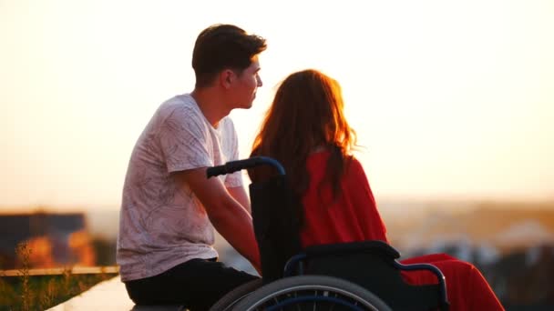 Mädchen mit roten Haaren sitzt im Rollstuhl und Freund sitzt bei ihr, sie reden über etwas und schauen in die Ferne - Filmmaterial, Video