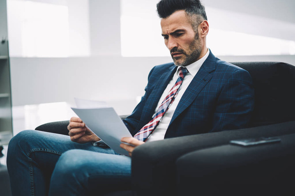 Concentrated experienced mature proud ceo dressed in formal wear reading accounting reports on papers sitting on armchair in office.Pensive businessman 50 years old checking financial information - Foto, afbeelding