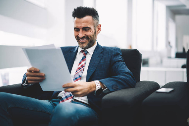 Mature successful businessman dressed in formal wear reading funny information on paper documents sitting on armchair in office interior.Cheerful proud ceo middle aged laughing during checking reports - Fotó, kép