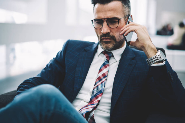 Confident mature entrepreneur in formal wear talking with business partner on smartphone and discussing strategy during phone call.Experienced proud ceo solving working issues on telephone device - Foto, Imagen