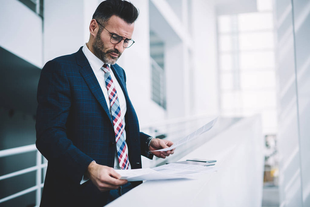 Concentrated smart businessman dressed in suit attentively reading financial text documents.Pensive proud ceo dressed in formal wear analyzing text reports with information data working in office - Fotografie, Obrázek
