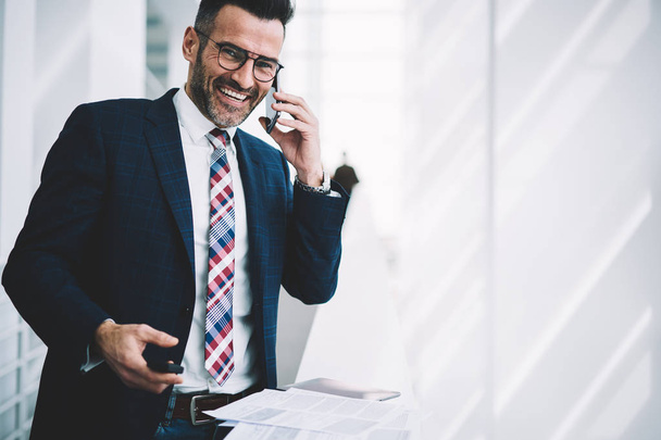 Portrait of successful businessman dressed in formal wear calling on smartphone device and laughing during phone conversation standing in office of corporate company near copy space area - Фото, изображение