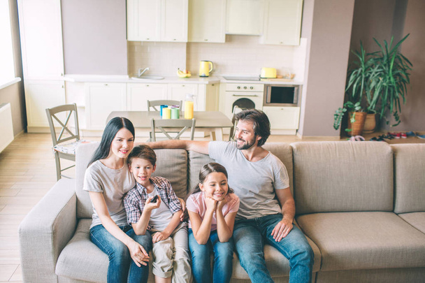 Beautiful family is sitting on sofa in kitchen. They spend time together. Guy smiles and looks at wife. Boy is leaning to mom. Girl sits with dad. They are looking straight forward with excitement. - Photo, image
