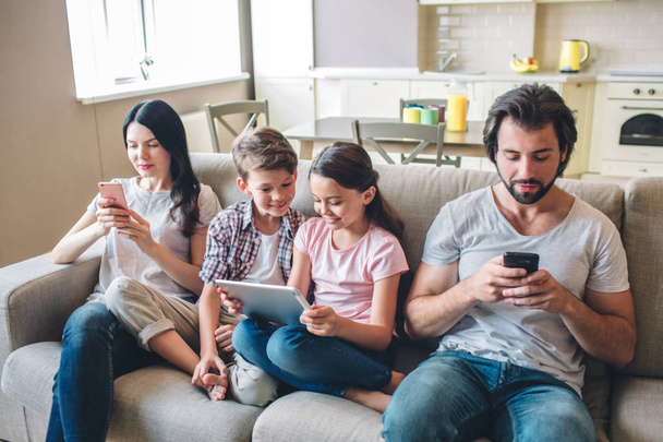 Parents are sitting on sofa with kids and look at phones. Children are in bettween woman and man. Girl holds tablet in hands. They look at screen. - Foto, afbeelding