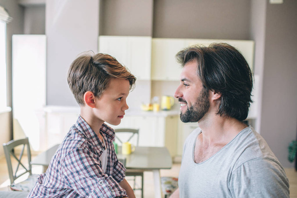 Happy man is looking at his son. He smiles. Boy looks at his dad with serious sight. They are alone in kitchen. - Фото, изображение