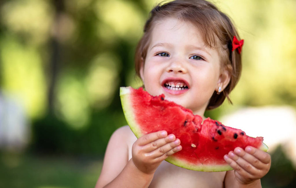 Grappige meisje bijten een segment van watermeloen buiten op warme en zonnige zomerdag. Kopiëren, lege ruimte voor tekst - Foto, afbeelding