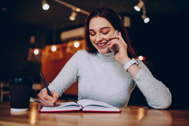 Smiling teen hipster girl making telephone call for sharing good news about completed coursework, happy young woman writing homework and talking on smartphone with friend satisfied with tariffs - Φωτογραφία, εικόνα