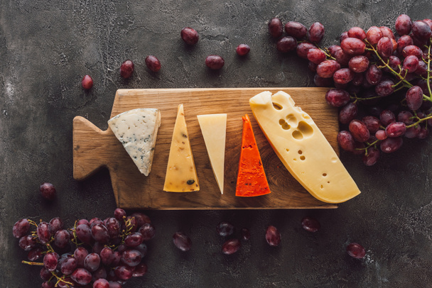 top view of assorted cheese on wooden cutting board with grape on dark surface - Photo, Image