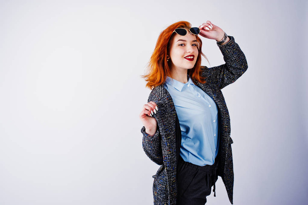 Portrait of a fantastic redheaded girl in blue shirt, grey overcoat posing with sunglasses in the studio. - Photo, Image