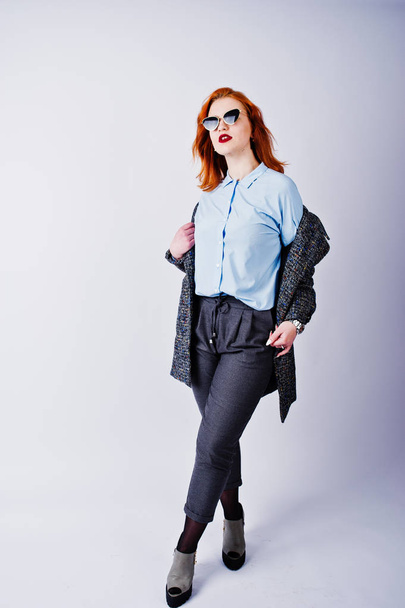Portrait of a fantastic redheaded girl in blue shirt, grey overcoat posing with sunglasses in the studio. - Fotografie, Obrázek