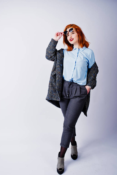 Portrait of a fantastic redheaded girl in blue shirt, grey overcoat posing with sunglasses in the studio. - Foto, imagen