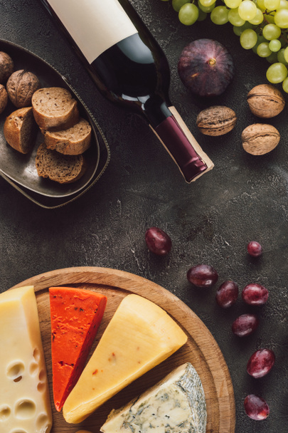food composition with bottle of wine, grape, bread pieces and assorted cheese on dark surface - Photo, Image