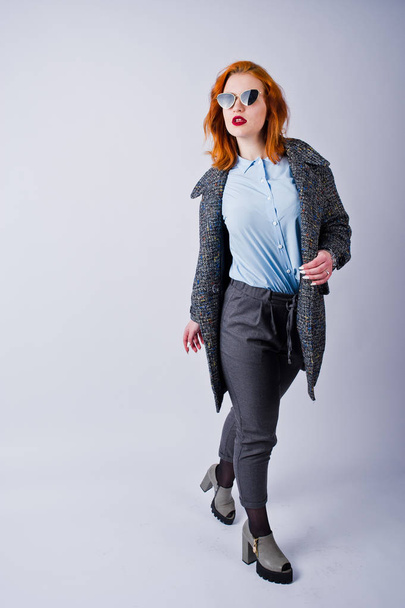 Portrait of a fantastic redheaded girl in blue shirt, grey overcoat posing with sunglasses in the studio. - Valokuva, kuva