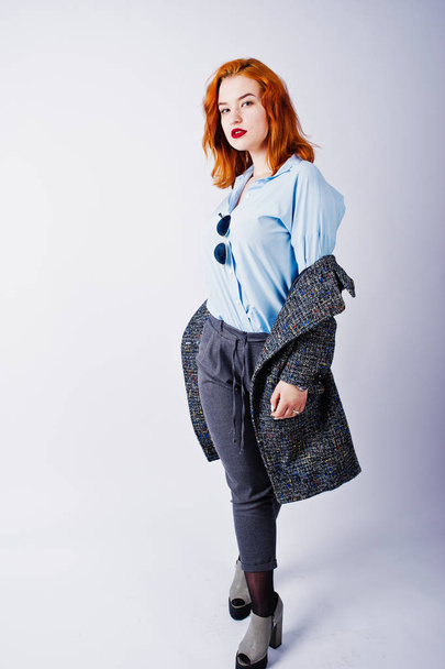 Portrait of a fantastic redheaded girl in blue shirt, grey overcoat posing with sunglasses in the studio. - Photo, image