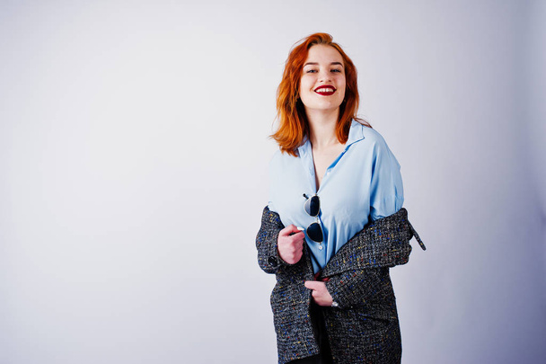 Portrait of a fantastic redheaded girl in blue shirt, grey overcoat posing with sunglasses in the studio. - Foto, afbeelding