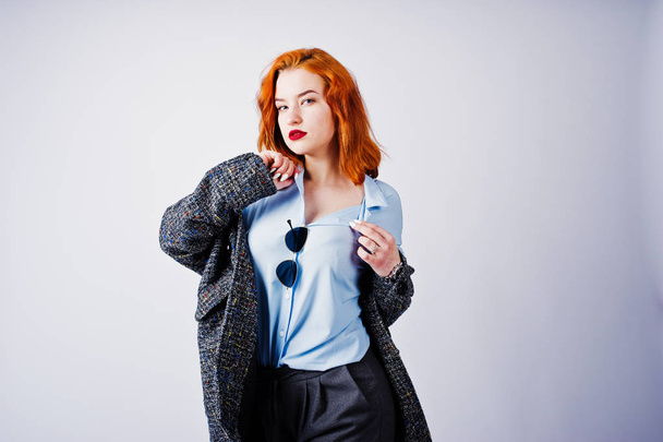 Portrait of a fantastic redheaded girl in blue shirt, grey overcoat posing with sunglasses in the studio. - Фото, зображення