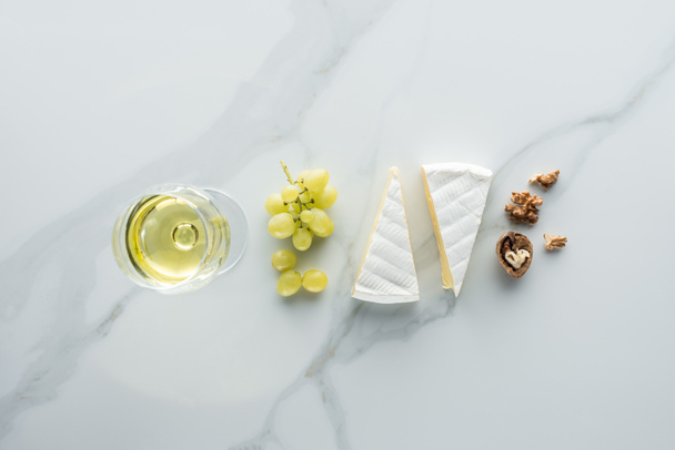 flat lay with glass of wine, camembert cheese and hazelnuts on white marble surface - Photo, Image