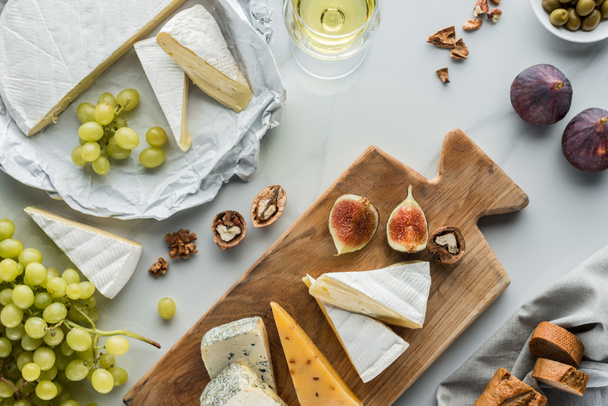 flat lay with food composition of cheese and fig pieces on cutting board, fruits and glass of wine on white marble surface - 写真・画像