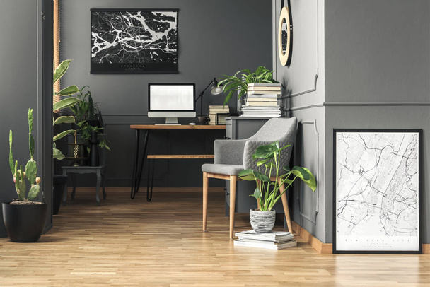 Fresh green plants in real photo of dark room interior with wainscoting on wall, grey armchair and wooden desk with empty screen computer - Foto, Imagen