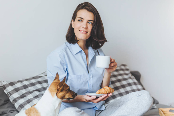 Porträt einer verträumten brünetten Frau sitzt mit überkreuzten Beinen auf einem bequemen Bett im Schlafzimmer, blickt nachdenklich zur Seite, hält eine Tasse Kaffee und Croissant und ihren Lieblingsjack Russell Terrier in der Hand - Foto, Bild