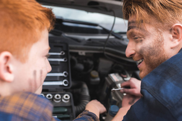 счастливый отец и сын ремонтируют машину с открытым капотом и смотрят друг на друга
 - Фото, изображение