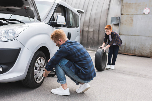 father changing tire in car with wheel wrench, son holding tire - Foto, immagini