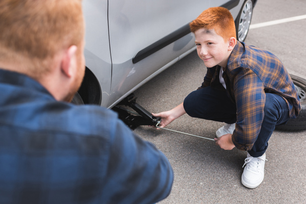 son lifting car with floor jack for changing tire and looking at father - Foto, Bild