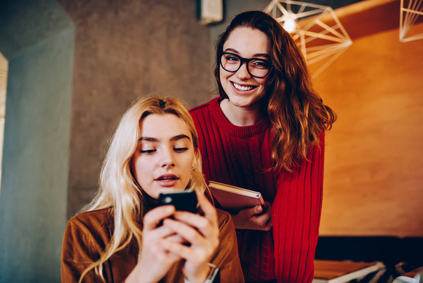 Portrait of cheerful teen woman spending time with friend which concentrated on sending text messages on smartphone, hipster girl networking via cellular ignoring communication with colleagu - Fotó, kép