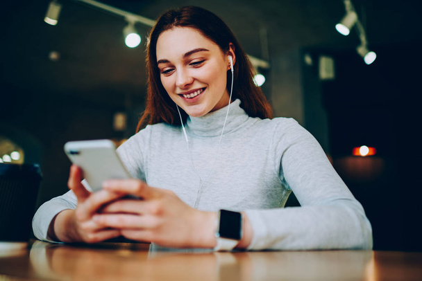 Cheerful teen girl watching favorite serial on smartphone via good wifi connection in cafe, smiling young woman using cellular for downloading new songs for playlist listening music on coffee brea - Zdjęcie, obraz