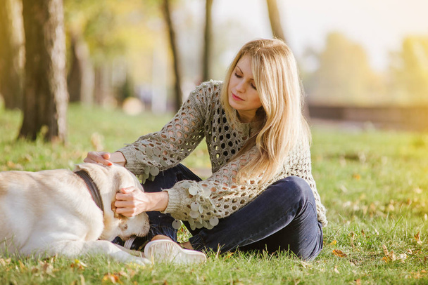 beautiful blonde is sitting on the grass in the park and playing with her dog labrador - Фото, зображення