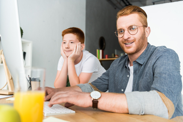 usměvaví otec a syn sedí u stolu s počítačem doma - Fotografie, Obrázek