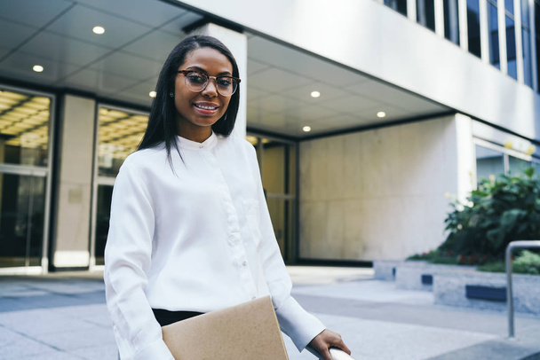 Portrait of successful african american female office employee dressed in white shirt holding folder with finance documents and smiling at camera standing in urban setting and smiling at camera - Foto, Imagem