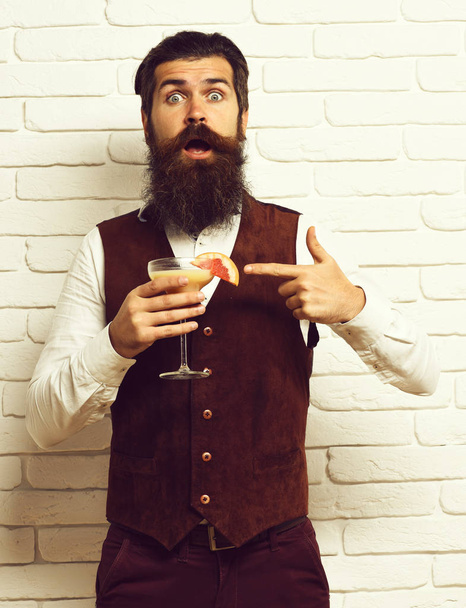 surprised face holding glass of alcoholic beverage in vintage suede leather waistcoat o white brick wall studio background. handsome bearded man with long beard and mustache has stylish hair on - Photo, Image